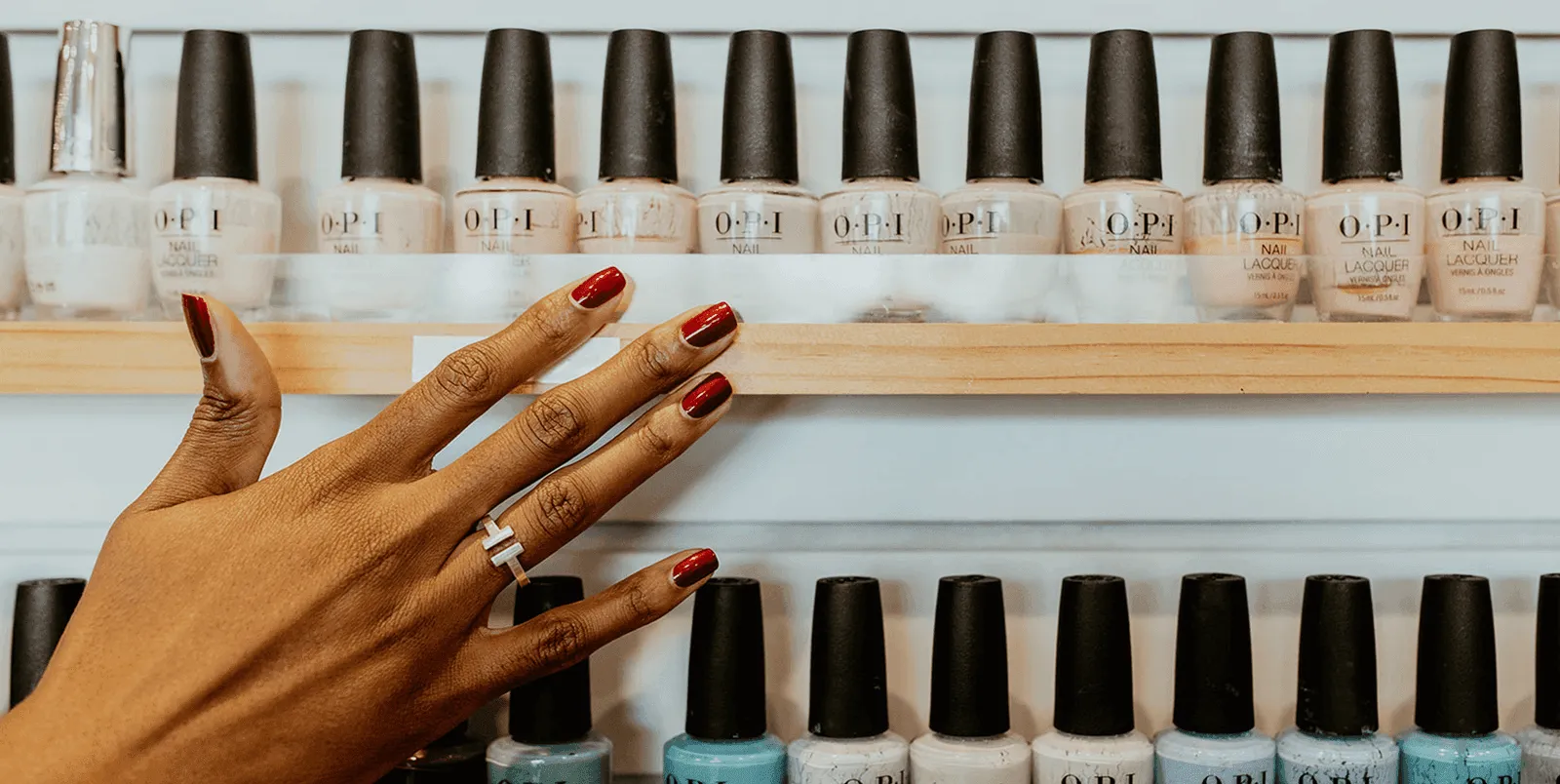A hand with a deep red manicure in front of shelves of premium nail polish
